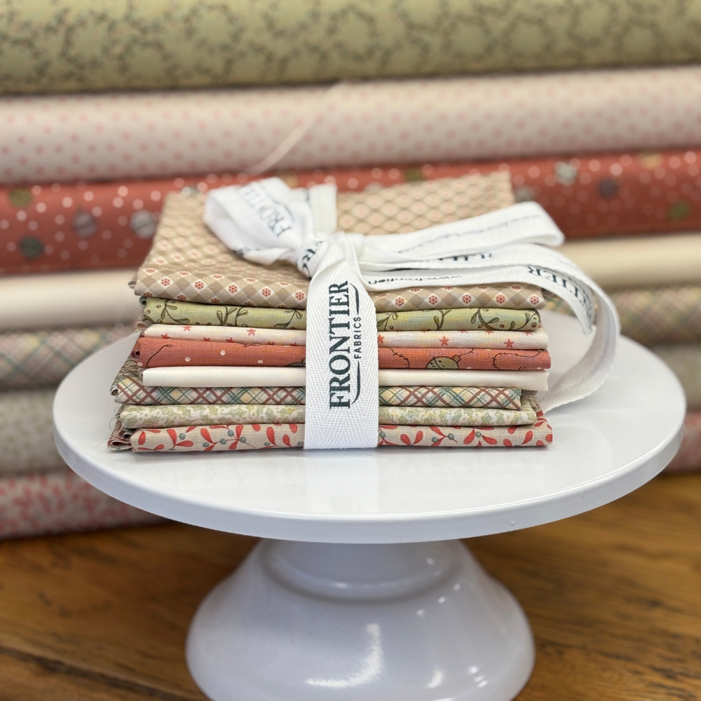 A bundle of Frontier Fabric fat quarters tied with a white ribbon, displayed on a white pedestal stand with a background of bolts of fabrics used in the bundle.
