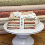 A bundle of Frontier Fabric fat quarters tied with a white ribbon, displayed on a white pedestal stand with a background of bolts of fabrics used in the bundle.