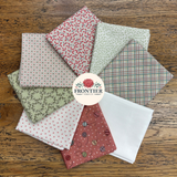 A set of Frontier Fabrics cotton fat quarters arranged in a fan shape on a rustic wooden background. The fabrics feature various patterns including plaid, polka dots, florals, and abstract prints in muted tones of red, green, pink, and white.