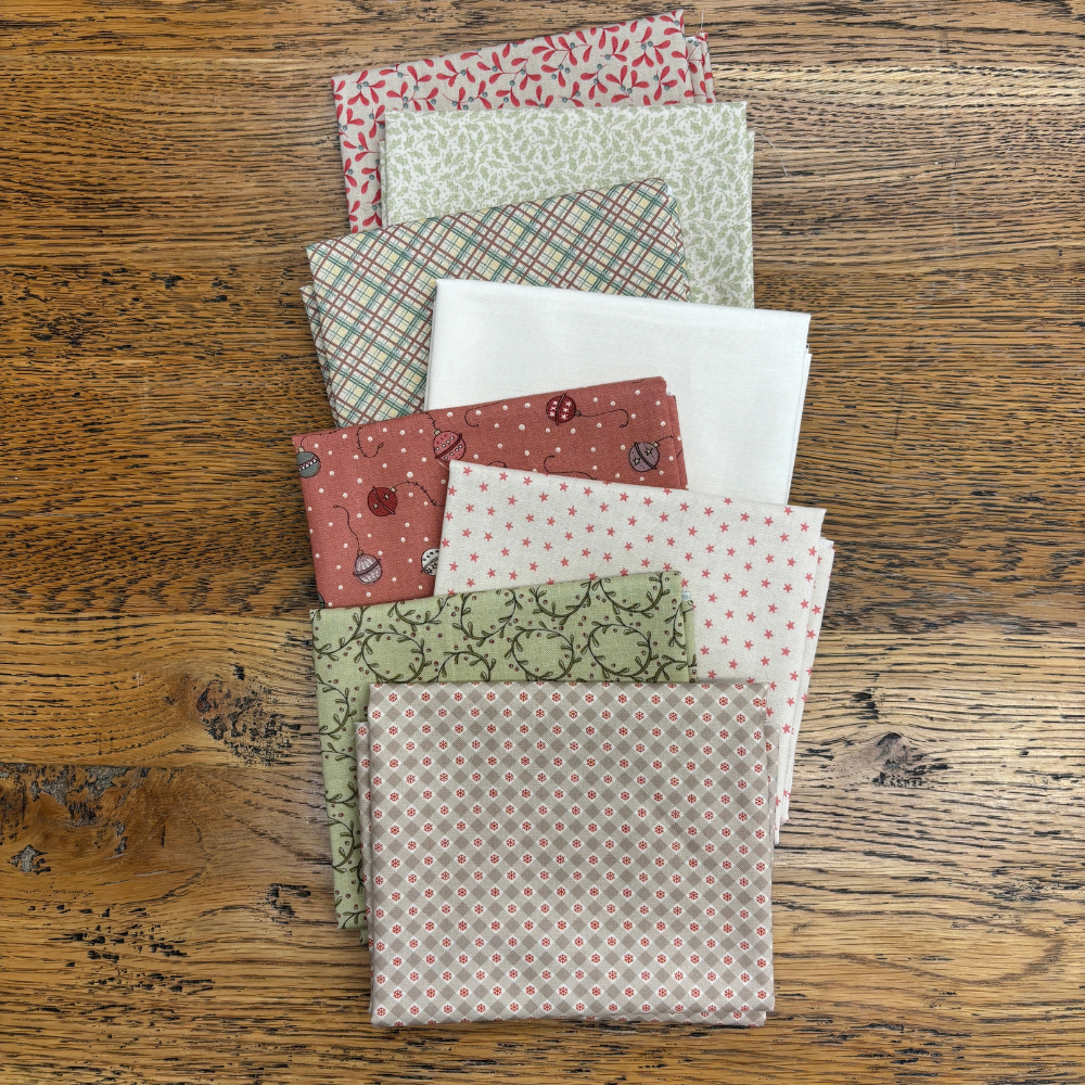 A stack of folded fabric fat quarters with various patterns and colours, including plaid, polka dots, and floral prints, arranged on a rustic wooden table.