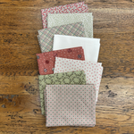 A stack of folded fabric fat quarters with various patterns and colours, including plaid, polka dots, and floral prints, arranged on a rustic wooden table.