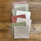 A stack of folded fabric fat quarters with various patterns and colours, including plaid, polka dots, and floral prints, arranged on a rustic wooden table.