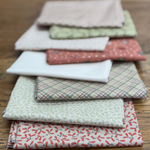 A stack of folded fat quarters in assorted colours and patterns, including floral, plaid, and solid designs, arranged on a wooden surface.