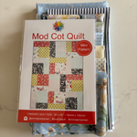 A packaged quilt pattern titled "Mod Cot Quilt" with a red "Mini Pattern" badge, placed on top of several folded quilt fabrics on a light surface.