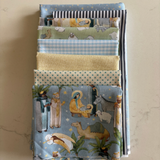 A stack of folded fabric pieces in various patterns and colours, including blue gingham, light blue with stars, butter yellow, and a blue nativity scene print, displayed on a marble surface.