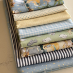 A stack of folded fabric pieces in assorted patterns and pastel colours, including blue gingham, cream polka dots, light green with animal prints, grey-and-white stripes, and starry blue fabric, photographed on a light neutral surface.