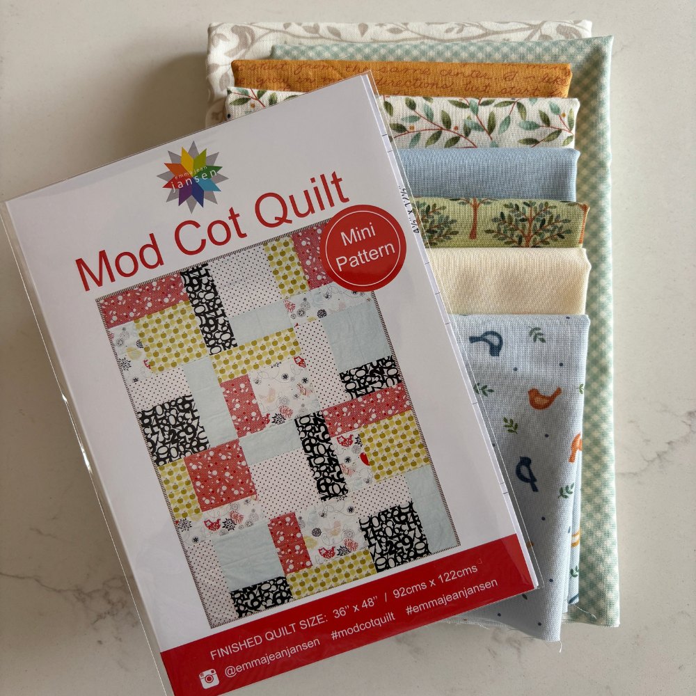 A packaged quilt pattern titled "Mod Cot Quilt" with a red "Mini Pattern" badge, placed over a stack of fabric swatches in assorted colours and patterns on a light stone surface.