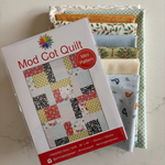 A packaged quilt pattern titled "Mod Cot Quilt" with a red "Mini Pattern" badge, placed over a stack of fabric swatches in assorted colours and patterns on a light stone surface.