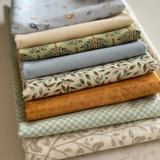 A stack of folded fabric pieces in soft pastel and earthy tones, including floral prints, gingham, and solid colours, arranged neatly on a light surface.
