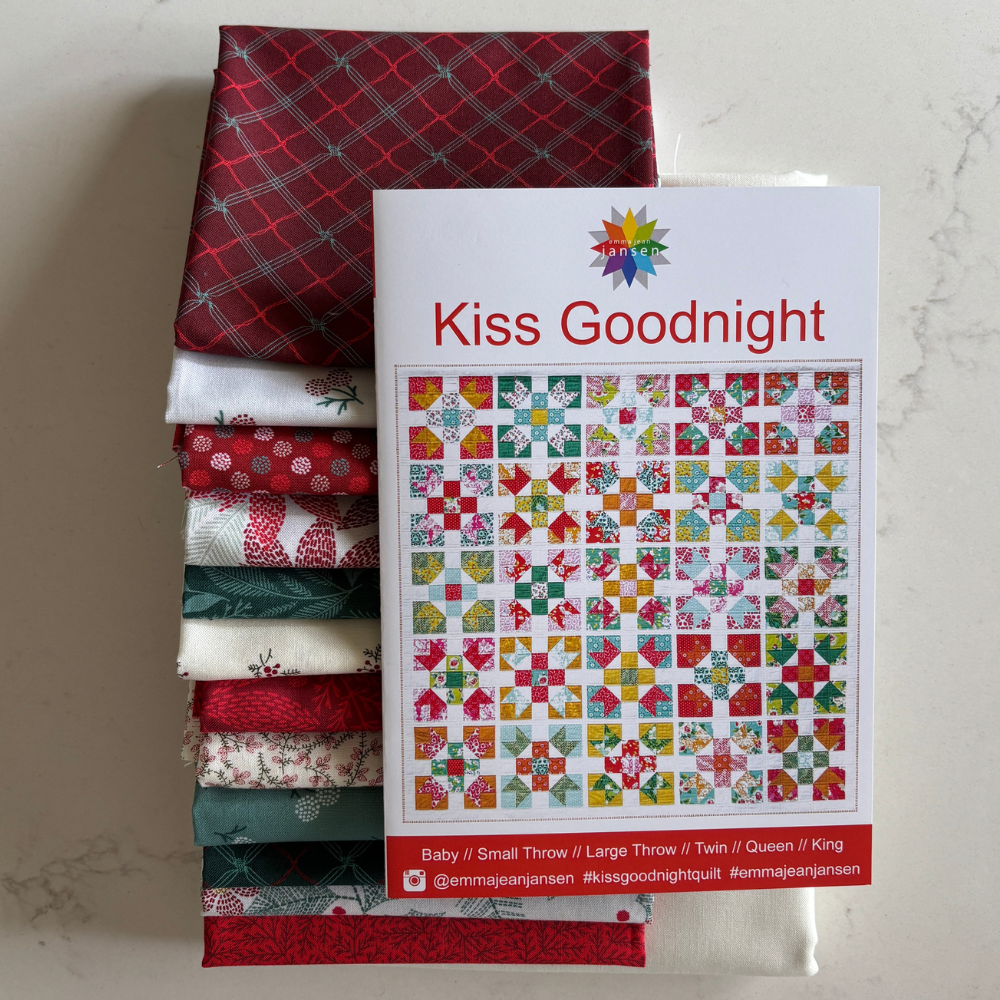 A stack of folded fat quarters in red, green, white, and patterned fabrics sits next to a printed quilt pattern cover titled 'Kiss Goodnight' by Emma Jean Jansen. The quilts and pattern are arranged on a light marble-like surface.