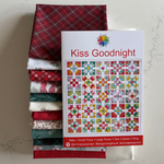 A stack of folded fat quarters in red, green, white, and patterned fabrics sits next to a printed quilt pattern cover titled 'Kiss Goodnight' by Emma Jean Jansen. The quilts and pattern are arranged on a light marble-like surface.