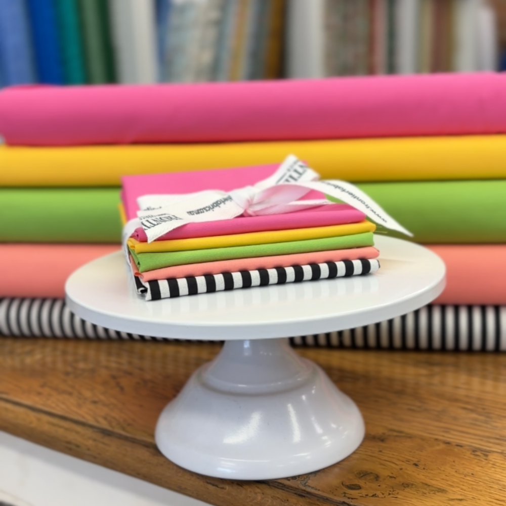 A stack of colourful fat quarters tied with a white ribbon sits on a white pedestal cake stand, with more fabric bolts and a blurred shop image in the background.