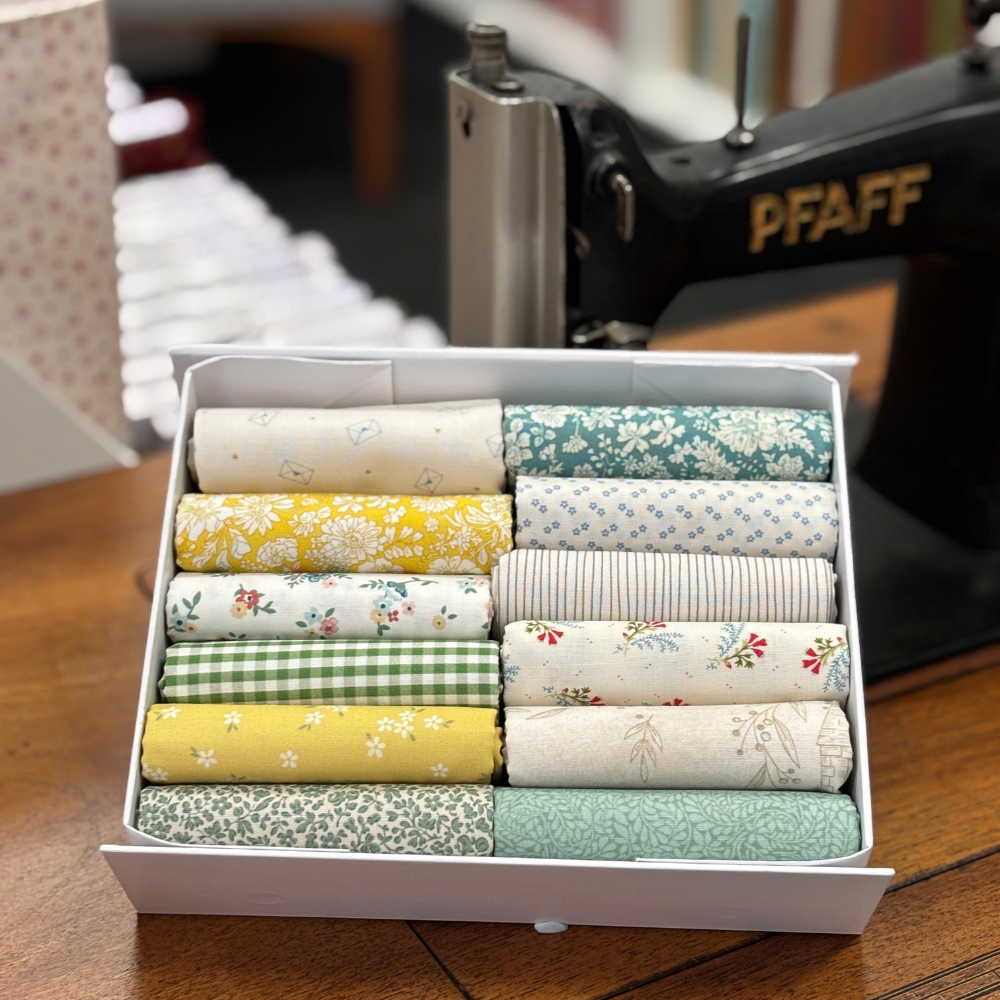 A white box filled with neatly folded fat quarters in various floral, striped, and gingham patterns, set on a wooden table with a PFAFF vintage sewing machine blurred in the background.