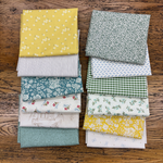 Two rows of folder fat quarters in various patterns (floral, gingham, stripes, polka dots) in green, yellow, blue, and white, arranged on a rustic wooden background.