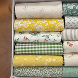 A close up image of folded fat quarters in a white box, showing various patterns and colours including teal floral, yellow floral, blue dot, green gingham, and beige botanical prints.