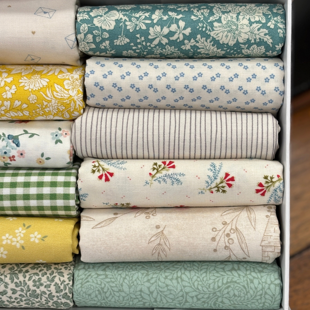 A close up image of folded fat quarters in a white box, showing various patterns and colours including teal floral, yellow floral, blue dot, green gingham, and beige botanical prints.