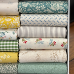 A close up image of folded fat quarters in a white box, showing various patterns and colours including teal floral, yellow floral, blue dot, green gingham, and beige botanical prints.