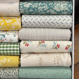 A close up image of folded fat quarters in a white box, showing various patterns and colours including teal floral, yellow floral, blue dot, green gingham, and beige botanical prints.