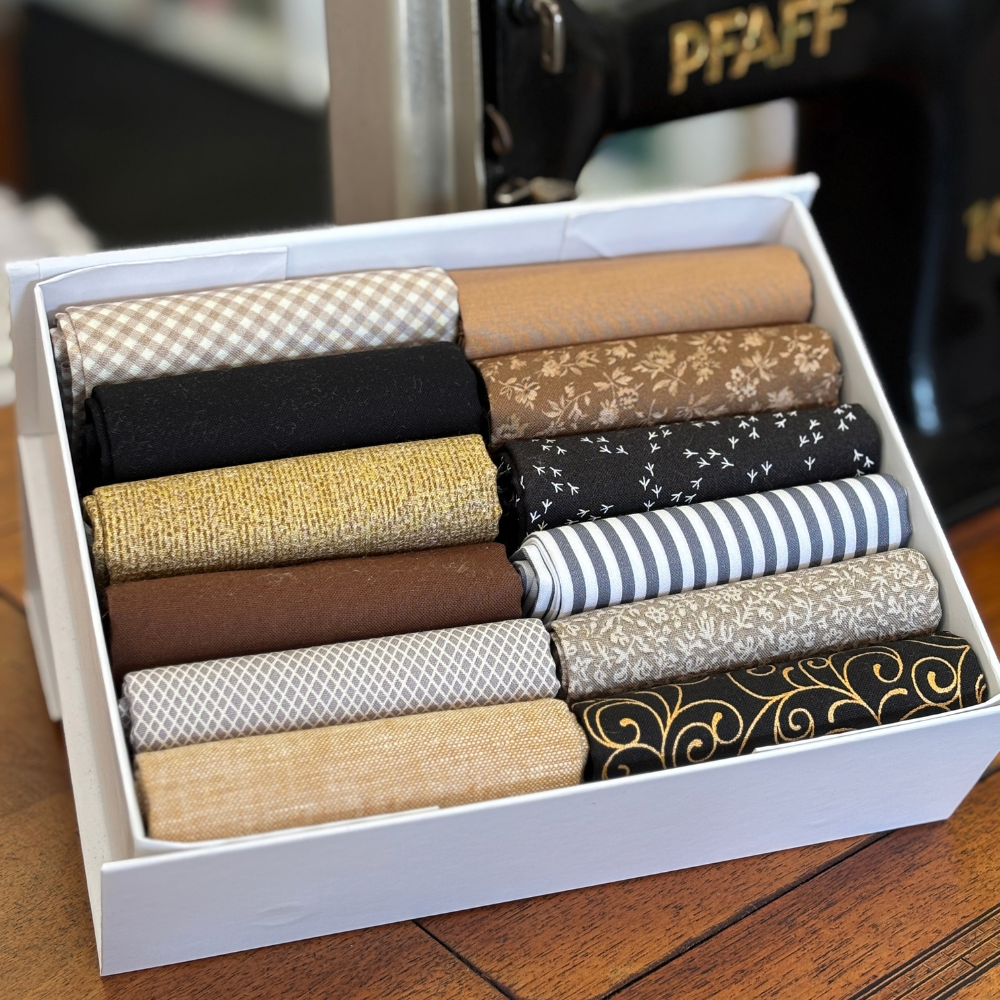 A white box filled with neatly fat quarters in assorted patterns and earthy colours, photographed on a wooden surface with a sewing machine in the background.