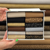 A white box filled with folded fat quarters in various patterns and colours, held open by two hands against a background of stacked fabric rolls.