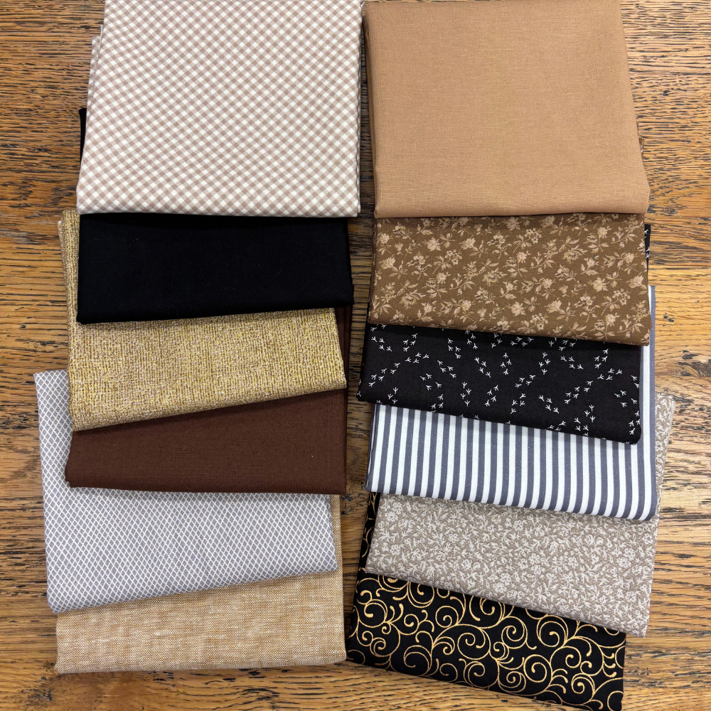 A stack of folded fat quarters with various patterns and colours, including gingham, floral, stripes, and metallic textures, arranged on a wooden surface.