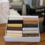 A white fabric box filled with neatly folded fat quarter fabrics in earthy tones, placed on a wooden table beside a vintage black sewing machine.