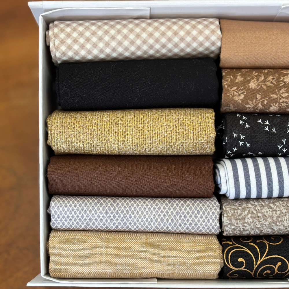 A white box filled with neatly folded fabric fat quarters in various patterns and colours, including beige gingham, black, floral brown, mustard yellow, dark grey with white birds, brown, navy and white stripes, light grey geometric, beige floral, and black with gold scrollwork.