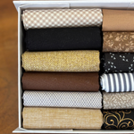 A white box filled with neatly folded fabric fat quarters in various patterns and colours, including beige gingham, black, floral brown, mustard yellow, dark grey with white birds, brown, navy and white stripes, light grey geometric, beige floral, and black with gold scrollwork.