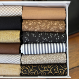 A box of folded fat quarters in assorted patterns and colorus, including beige gingham, solid tan, floral brown, black with white birds, striped blue and white, and black with gold scrollwork.