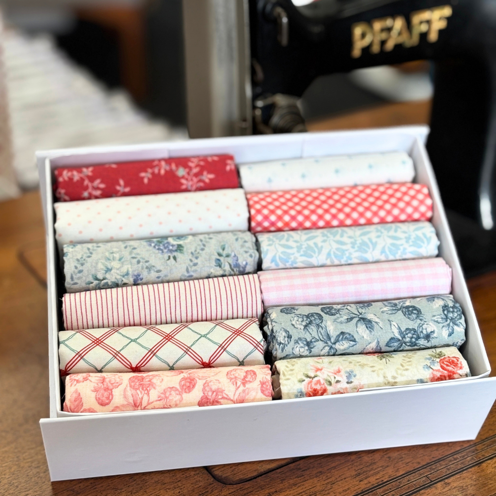 A white box filled with neatly folded fabric rolls in assorted floral, striped, gingham, and plaid patterns, photographed on a wooden surface with a sewing machine blurred in the background.