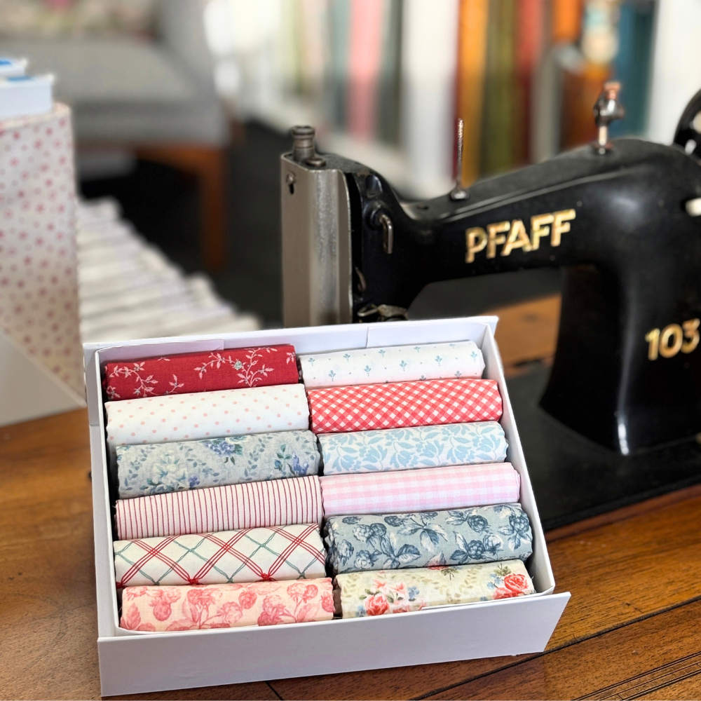 A white box filled with folded patterned quilting fabrics arranged in rows, placed on a wooden table beside a black PFAFF sewing machine. The fabrics feature various floral, gingham, striped, and plaid designs in red, pink, blue, green, and white.