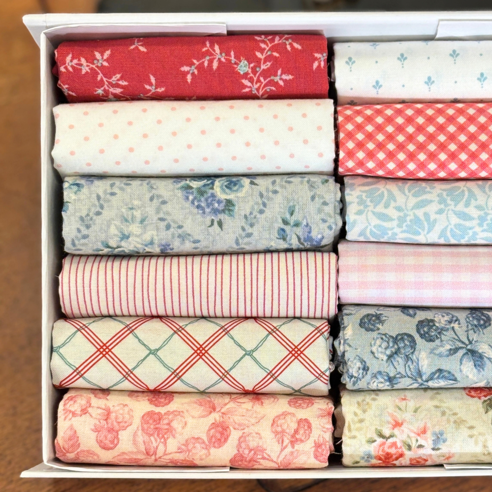 A white fabric storage box filled with neatly folded fat quarters in assorted patterns and colorus, including red floral, white polka dots, red gingham, blue floral, pink striped, and cream floral prints.