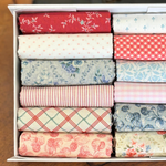 A white fabric storage box filled with neatly folded fat quarters in assorted patterns and colorus, including red floral, white polka dots, red gingham, blue floral, pink striped, and cream floral prints.