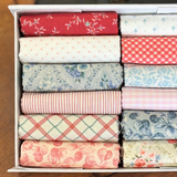 A white fabric storage box filled with neatly folded fat quarters in assorted patterns and colorus, including red floral, white polka dots, red gingham, blue floral, pink striped, and cream floral prints.