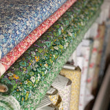 A close-up, angled view of multiple rolls of floral fabric stacked on shelves. The most prominent roll in the foreground features a green base with a dense yellow, white, and purple wildflower pattern, with other rolls in blue, pink, and yellow floral prints visible behind it.